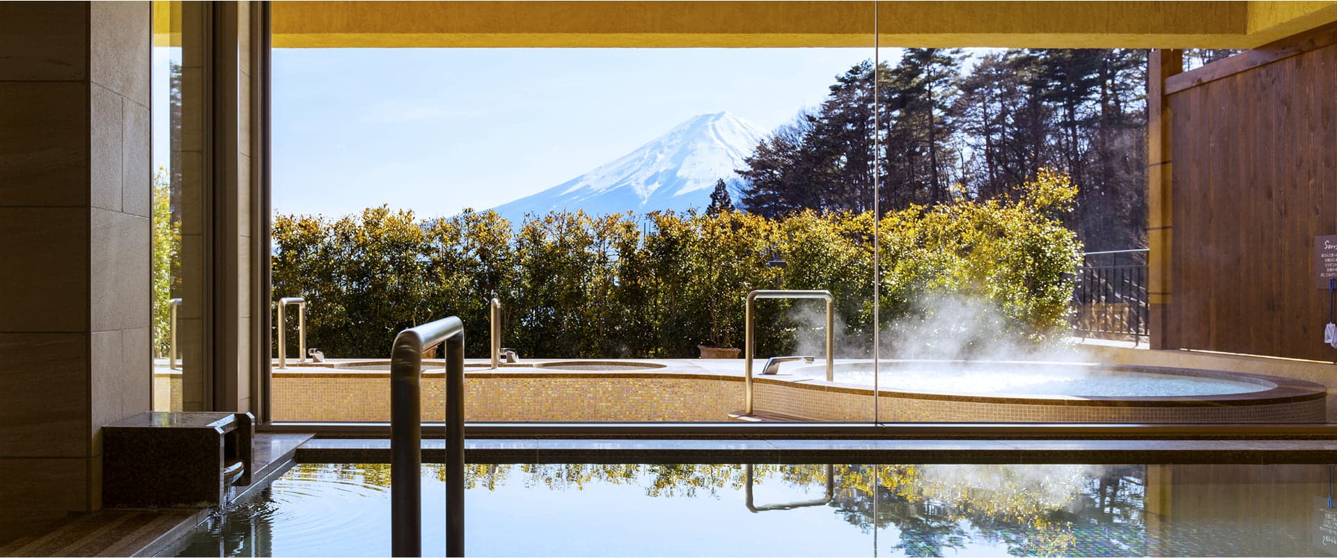 Open-air rotenburo onsen at La Vista Fuji Kawaguchiko with steam rising and a direct view of snow-capped Mt Fuji over the treeline