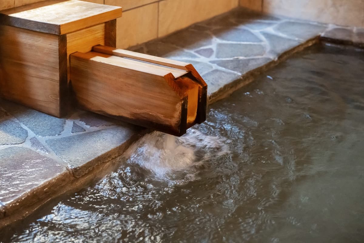 Close detail of a wooden spout pouring hot spring water into a stone-edged bath