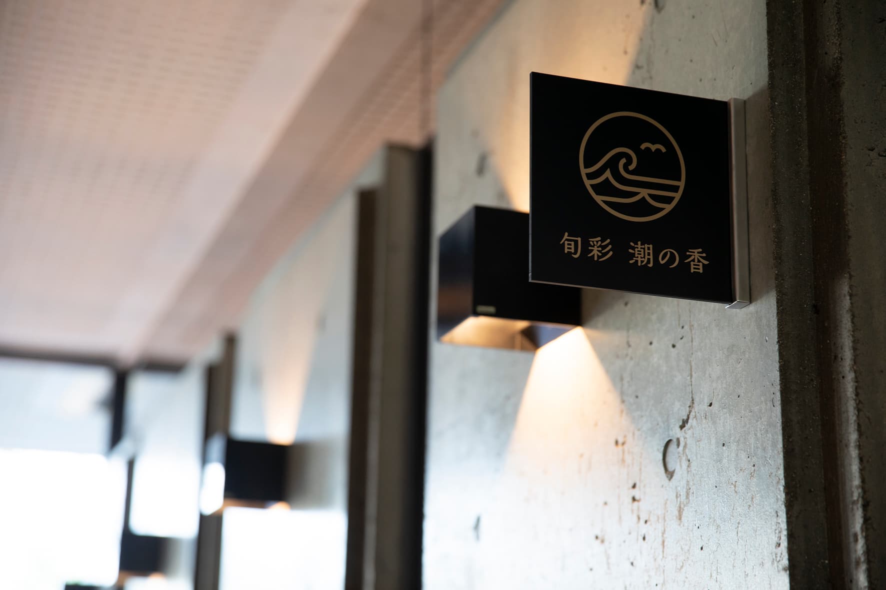 Concrete corridor at IBUKU lit by wall sconces leading to the in-house restaurant marked by a wave-motif plaque