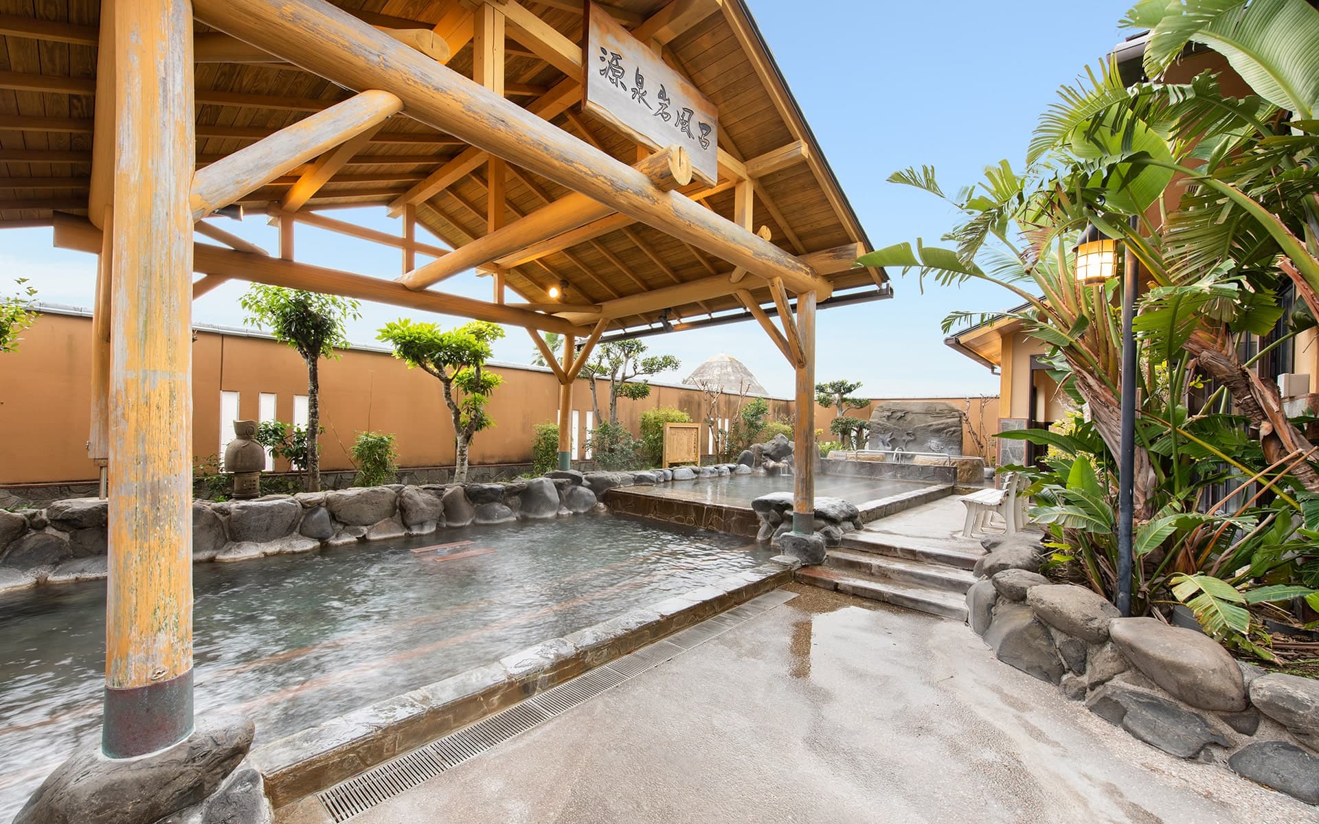 Open-air rotenburo hot-spring bath under a wooden pavilion surrounded by garden plantings and rising steam at Ibusuki Kokoro no Yado