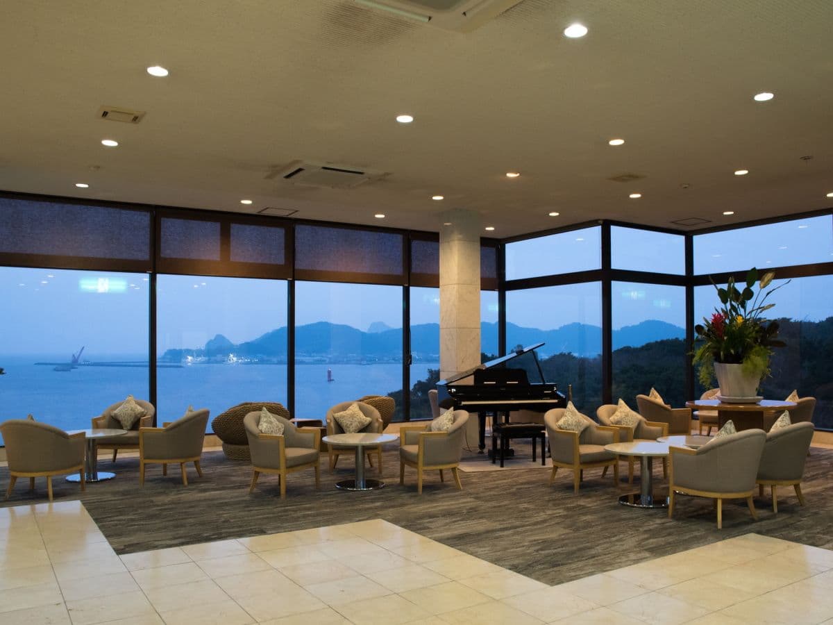Ocean-view lounge with armchairs and a grand piano framed by floor-to-ceiling windows at dusk