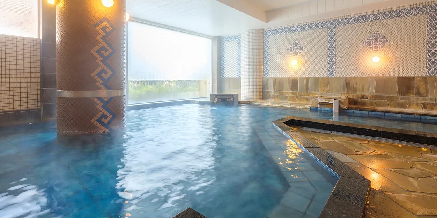 Large indoor public onsen bath at Kamenoi Hotel Atami with a mosaic-tiled pillar and blue thermal water
