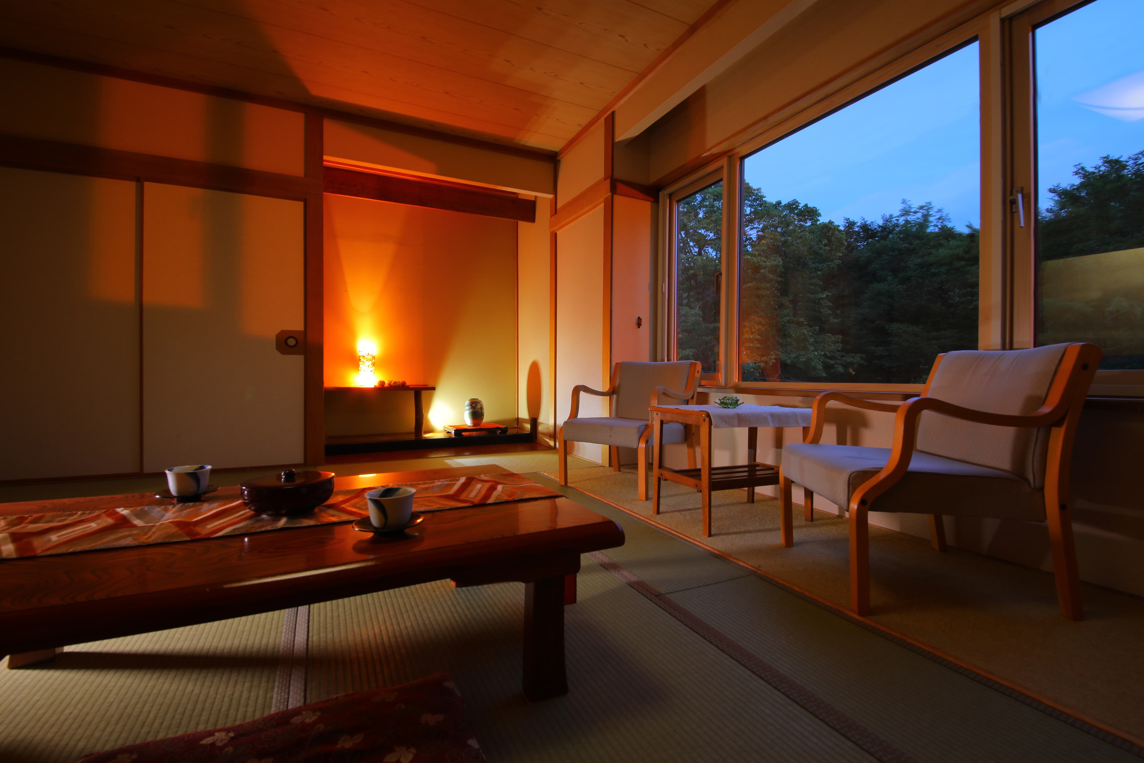 Tatami guest room at dusk with warm lighting, armchairs and a window framing the surrounding woods at Sanseikan