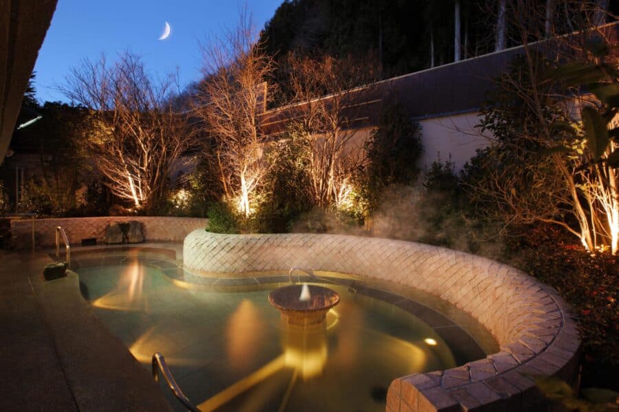 Outdoor rotenburo hot spring bath at dusk with steam, garden lighting and crescent moon at Yume no Toki