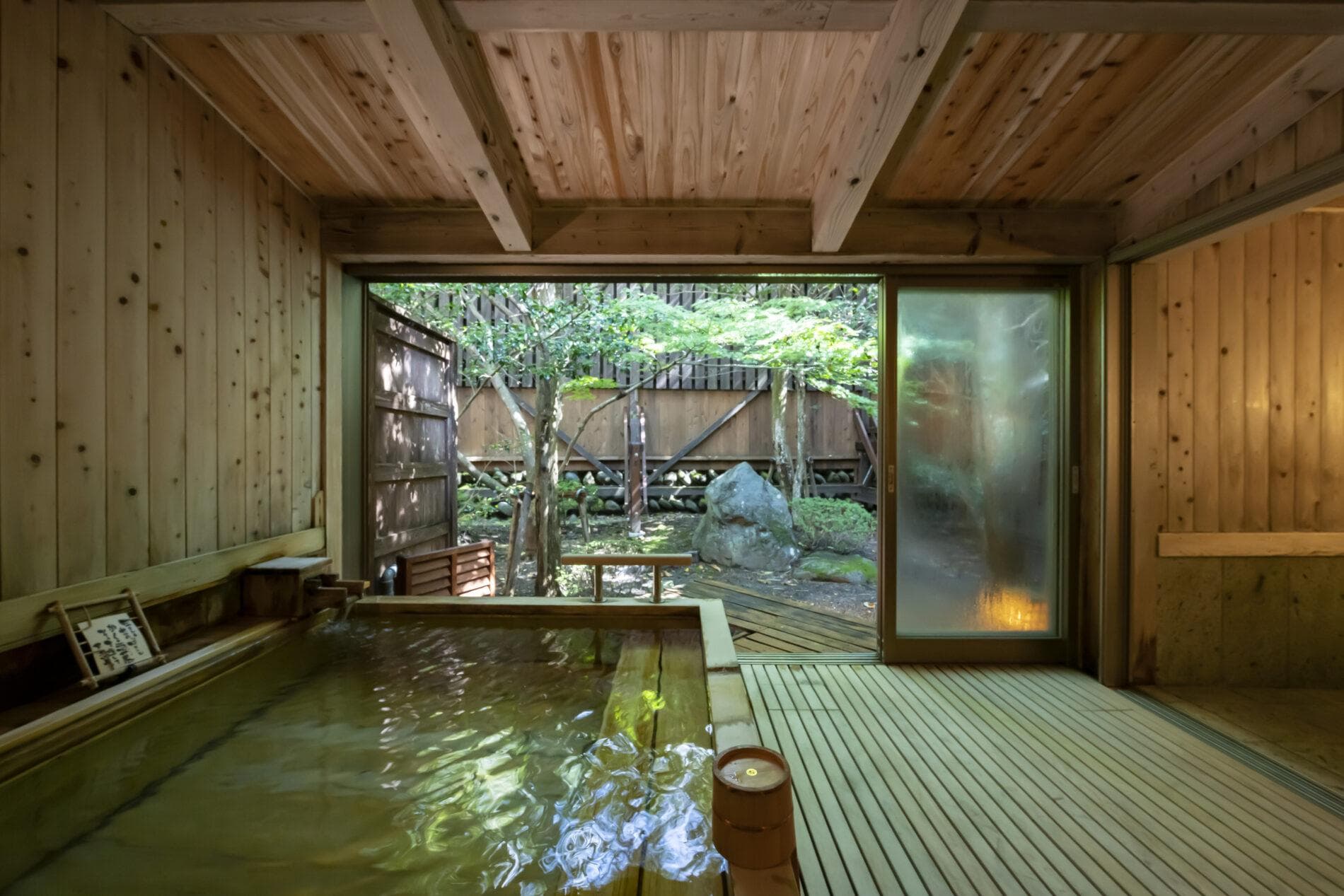 Private stone open-air bath opening onto a wooden deck and greenery