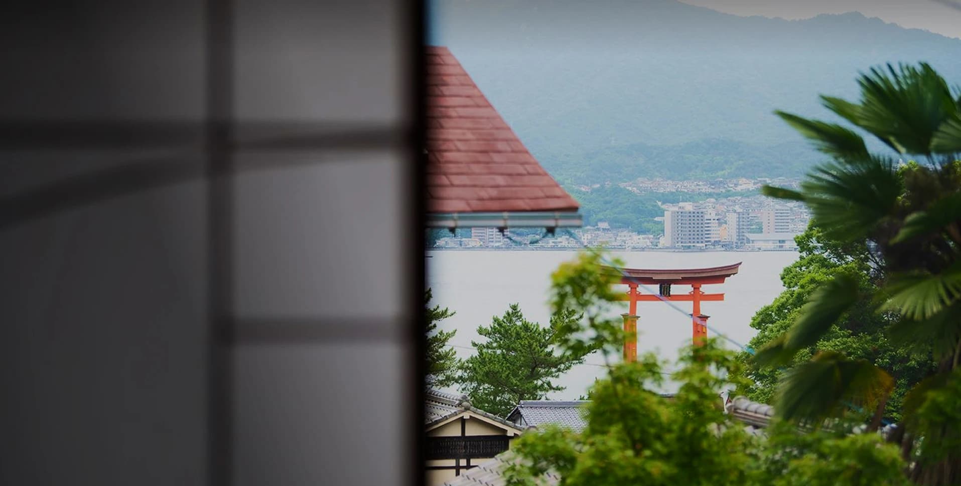 View of Itsukushima Shrine's vermilion floating torii gate seen through greenery from a Hotel Kikunoya window on Miyajima