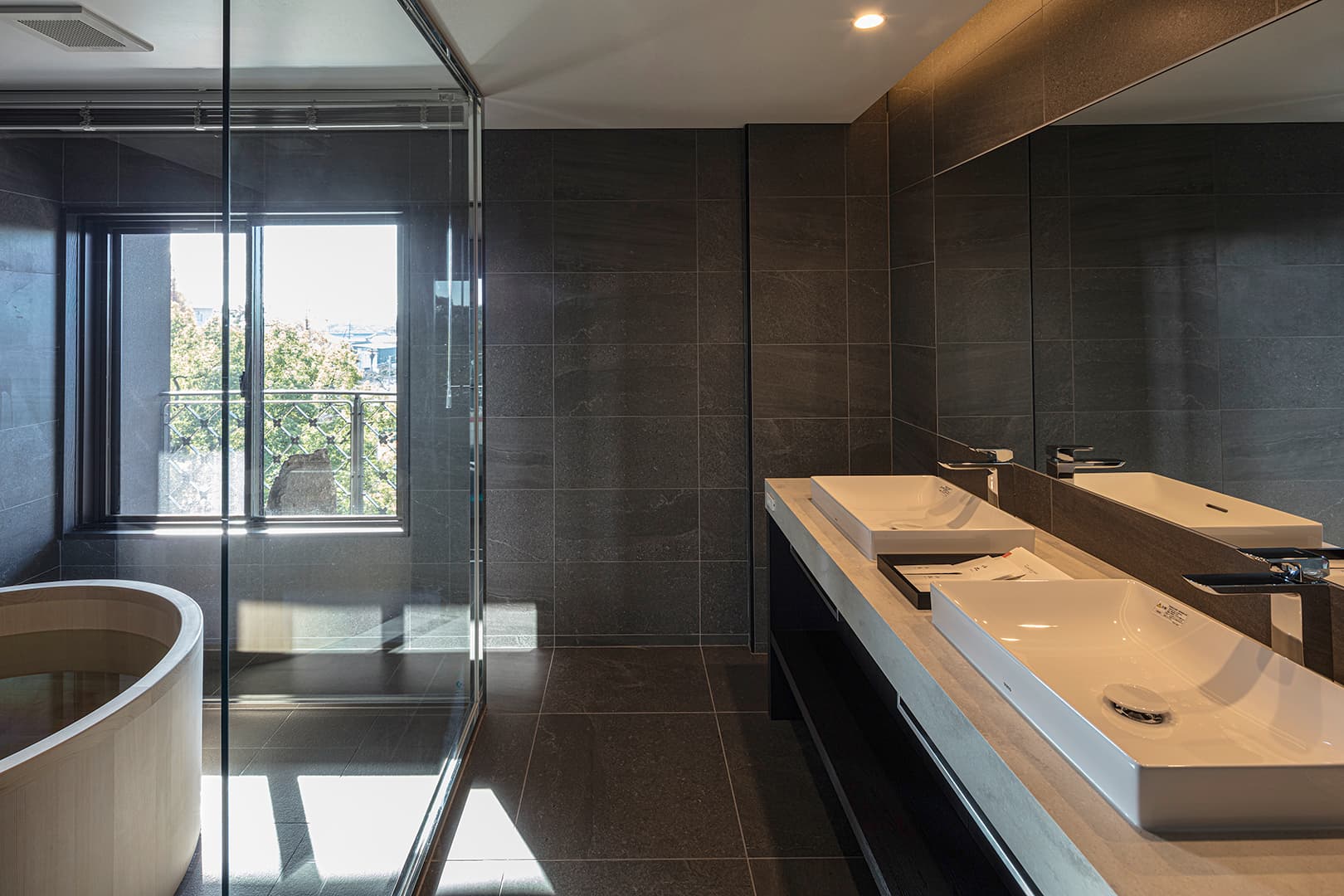 Modern stone-clad guest bathroom at Ohana with a wood-rimmed soaking tub, twin vessel sinks and a window over the Yanagawa townscape