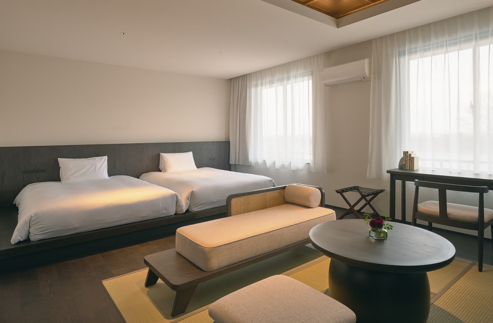 Spacious Ohana suite with twin beds, a cane daybench, round low table and sheer-curtained windows in warm afternoon light