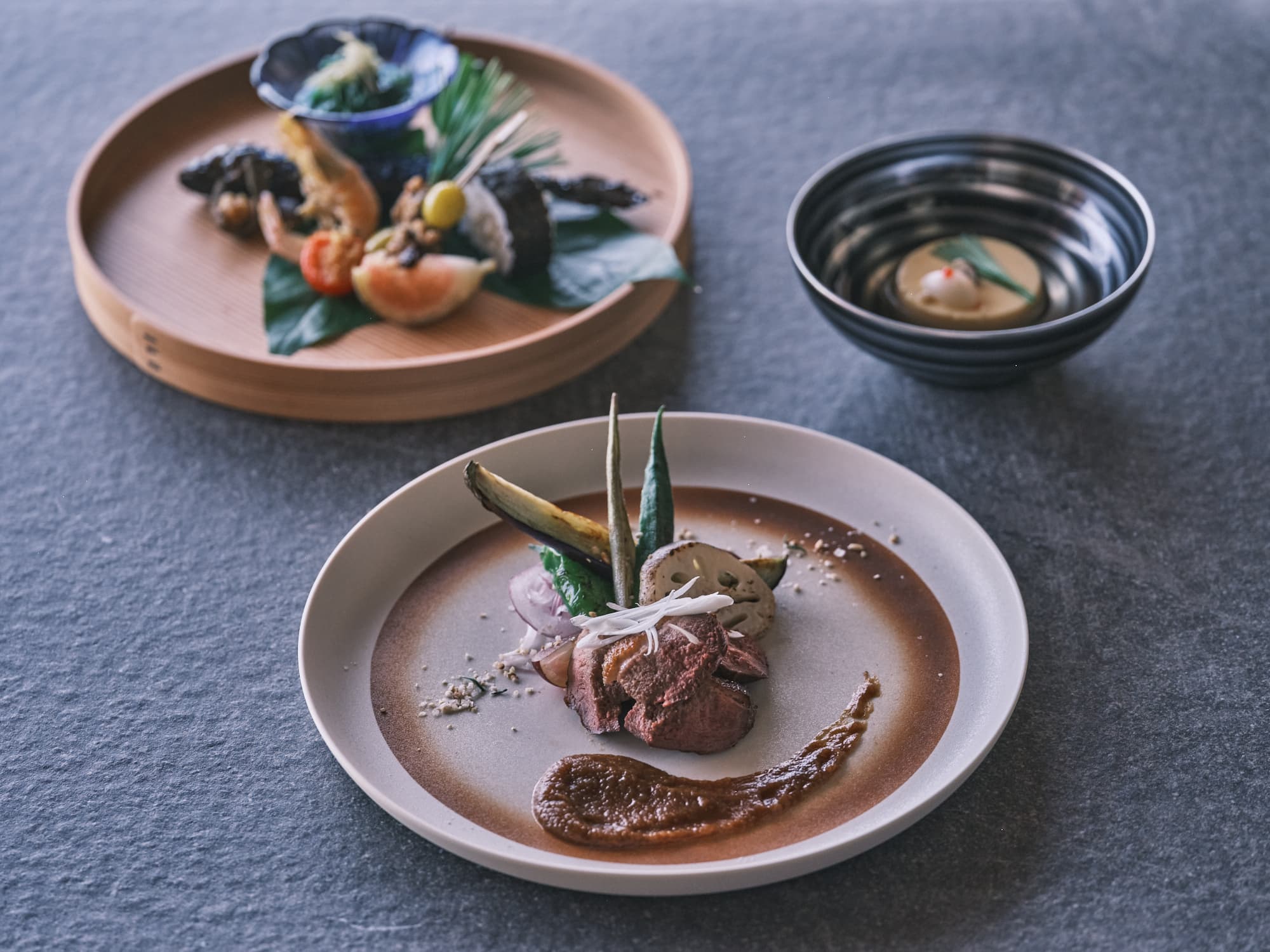 Ohana kaiseki dinner course of seasonal appetizers on a wooden tray and a plated meat dish with sauce on a stone tabletop