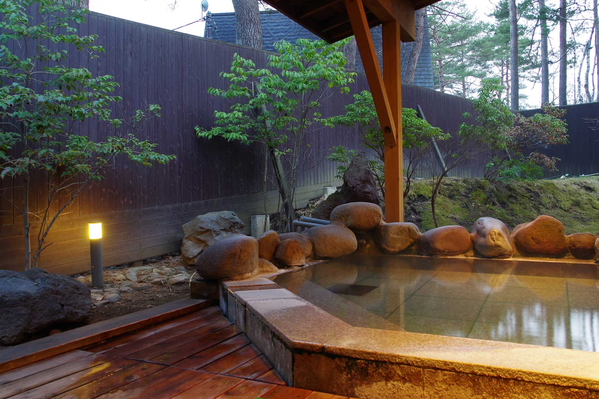 Open-air rotenburo at Kusatsu Skyland Hotel at dusk with a stone-edged pool surrounded by trees and a lantern