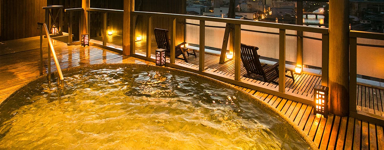 Rooftop open-air onsen bath glowing with lanterns at night beside wooden deck chairs at Honjin Hiranoya Kofukan