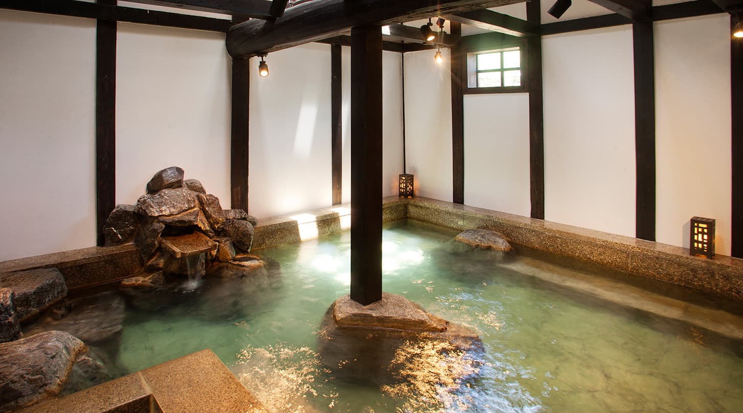 Indoor stone onsen bath with steam, rough granite edges and dark timber beams at Honjin Hiranoya Kofukan in Takayama