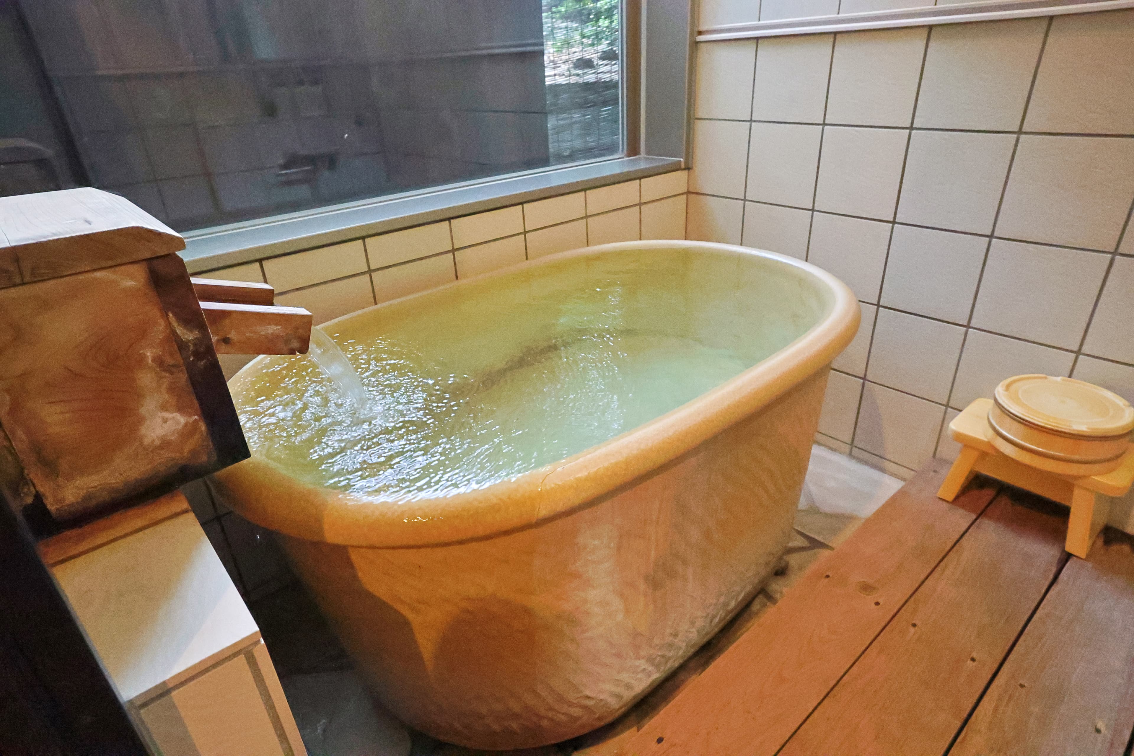 Private indoor Shigaraki-ware tub by a window with a wooden spout filling it with thermal water
