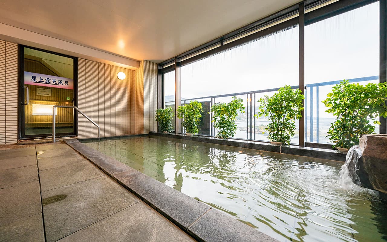 Indoor onsen bath with stone surround and large windows facing Atami bay