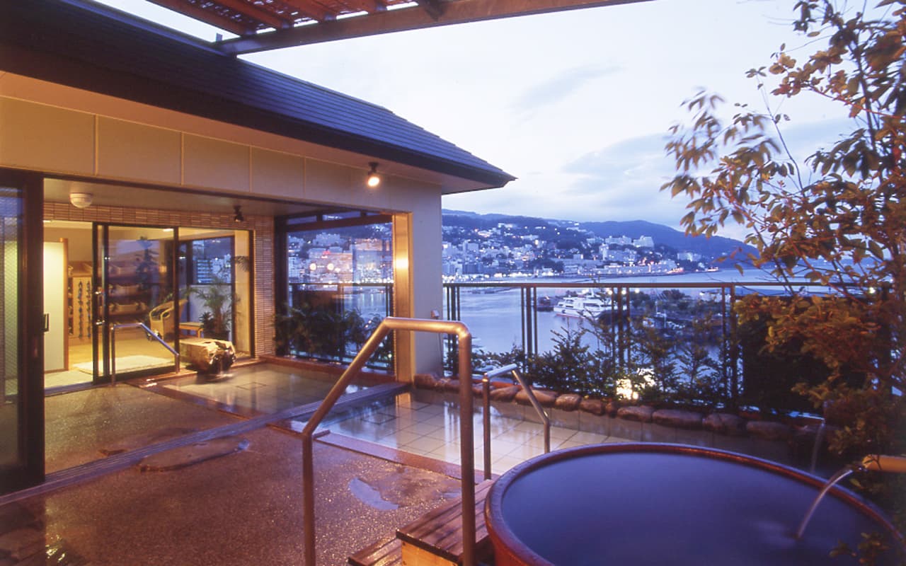 Sixth-floor Hana-no-yu open-air bath with ceramic jar tub and Atami harbour view at twilight