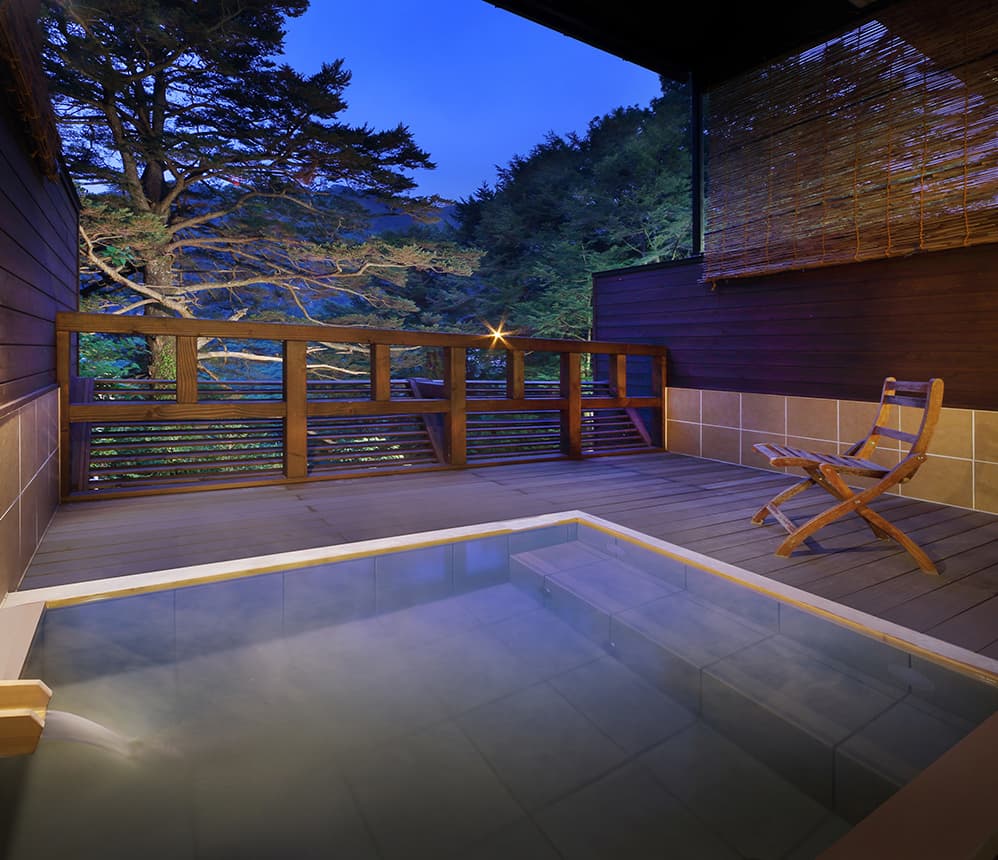 Private open-air onsen bath on a guest room terrace at dusk, framed by trees
