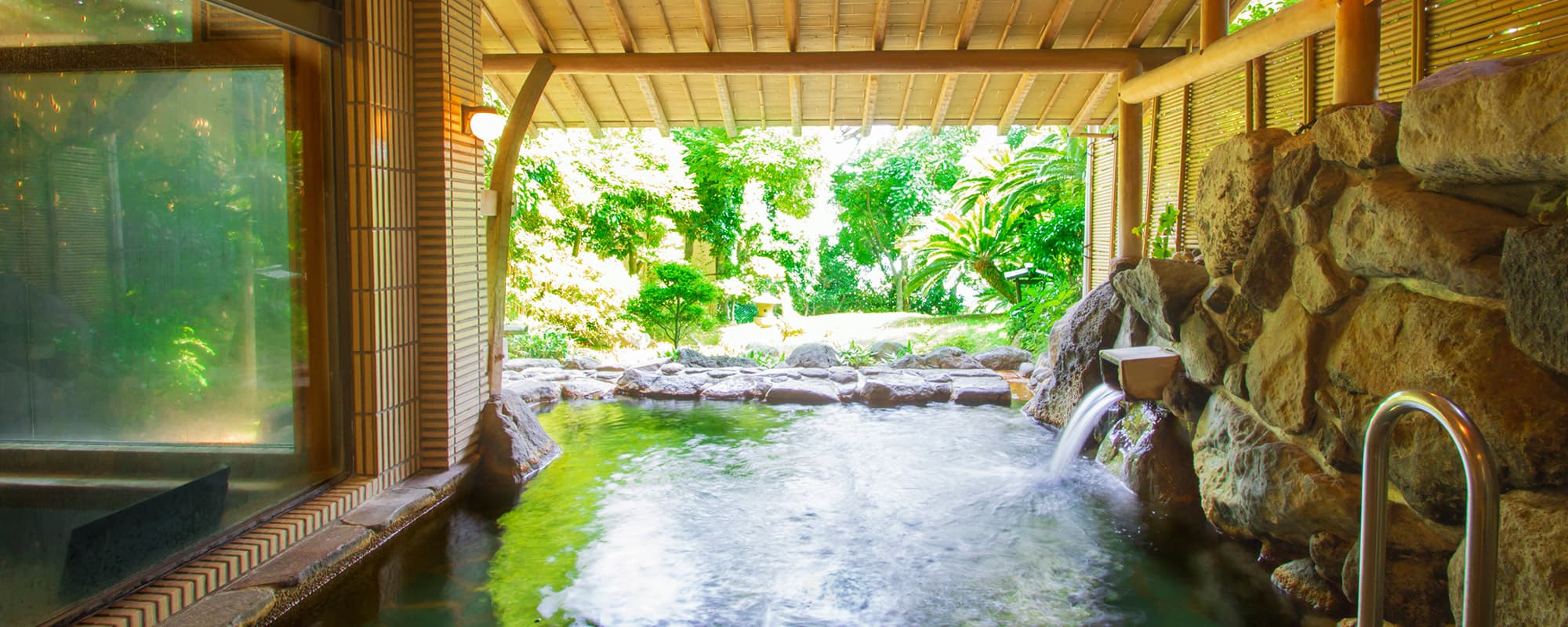 Outdoor onsen bath under a wooden roof opening onto a lush green garden at Atami Taikanso