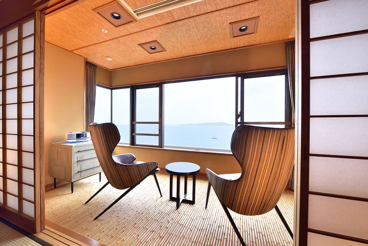 Corner sitting area of a sixth-floor Bousui guest room with two designer chairs facing a panoramic sea-view window