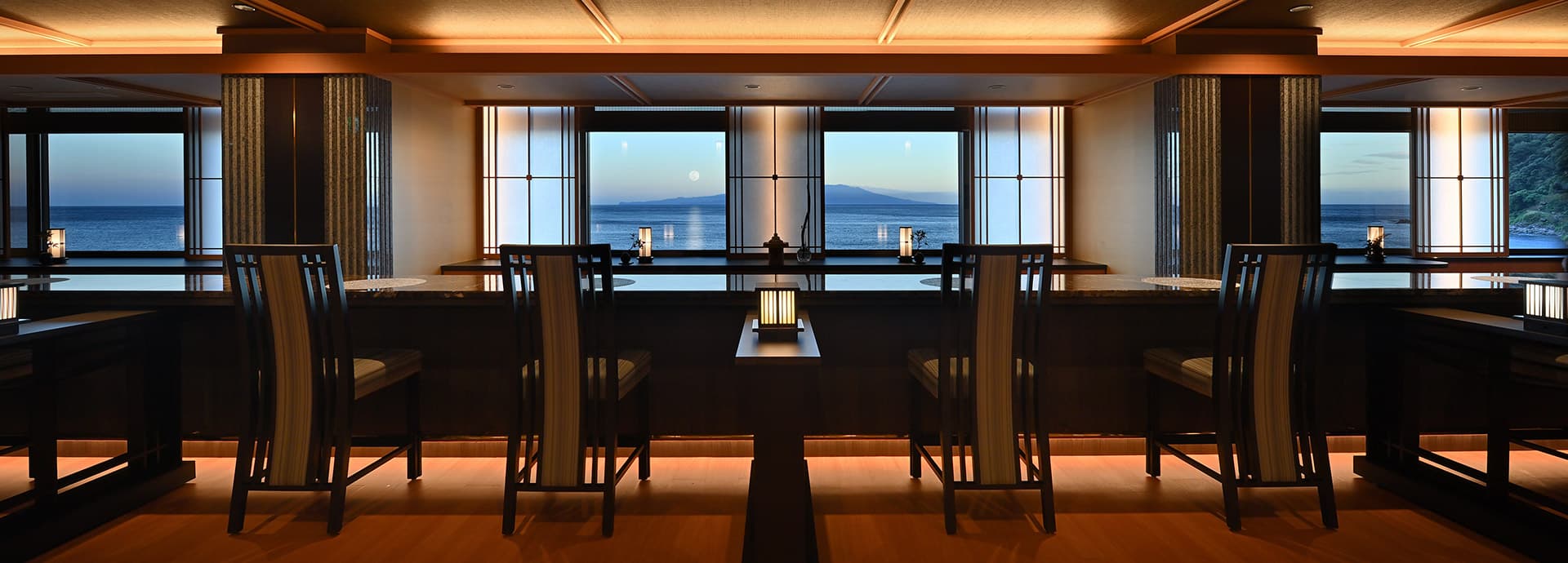 Sea-view lounge at Bousui at twilight with paired chairs along the windows and the moon rising over the bay