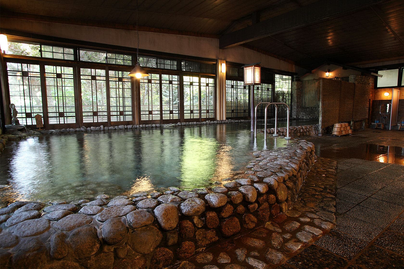 Spacious indoor hot spring bath at Daimaru Besso with a stone-edged pool and lattice windows framing the garden