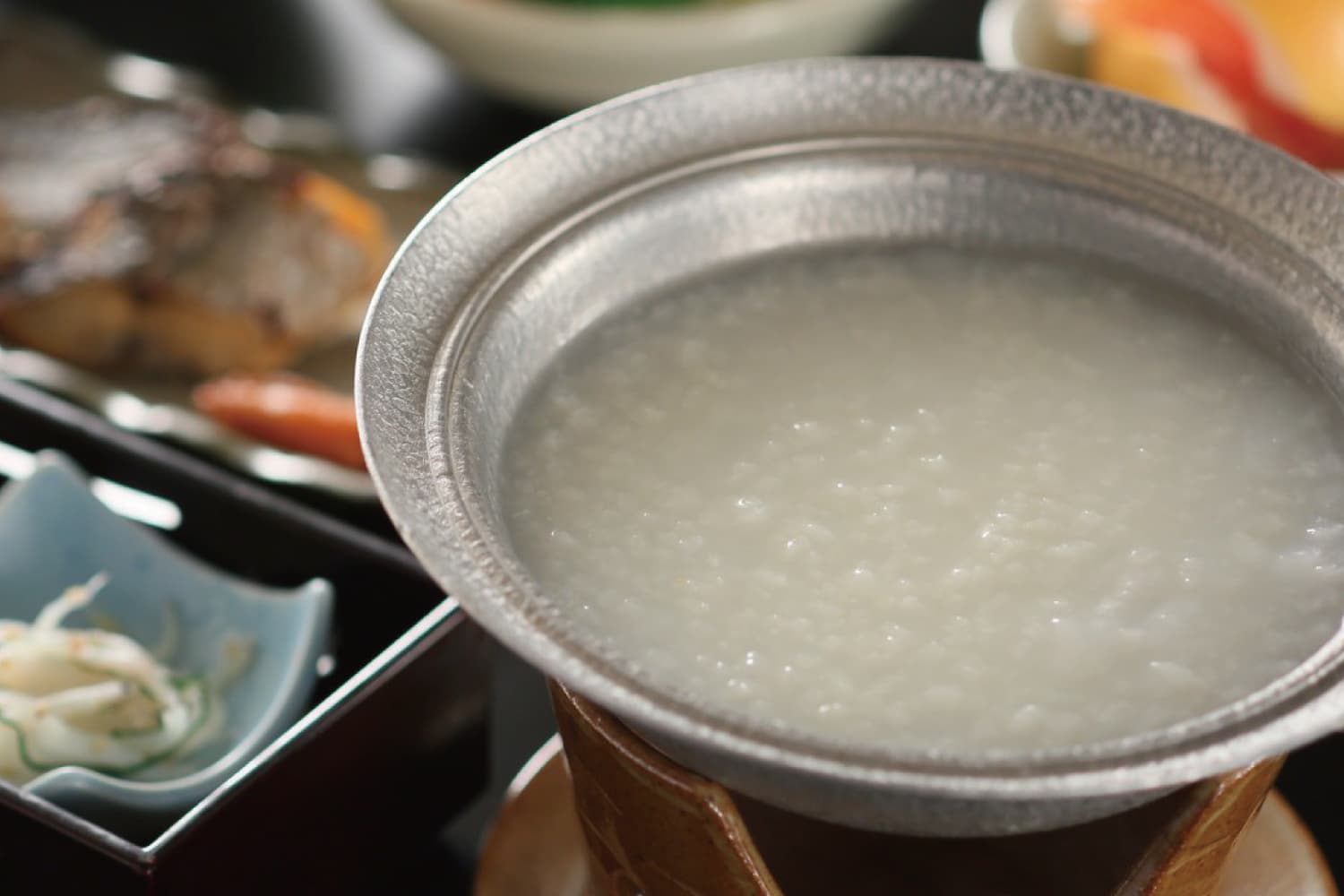 Signature hot-spring rice porridge simmering in a metal pot beside grilled fish at Shirahone Ebisuya