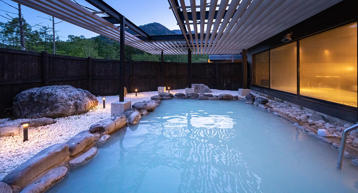 Outdoor rotenburo at dusk with milky Yumoto onsen water, stone lanterns and mountains beyond