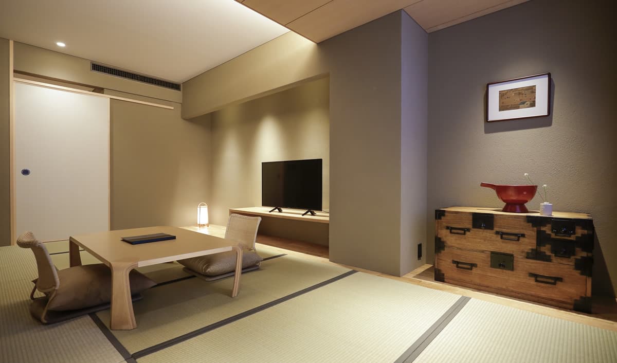 Warm-lit tatami guest room with antique tansu chest, framed artwork and floor seating at Kaminarimon Ryokan, Asakusa Tokyo