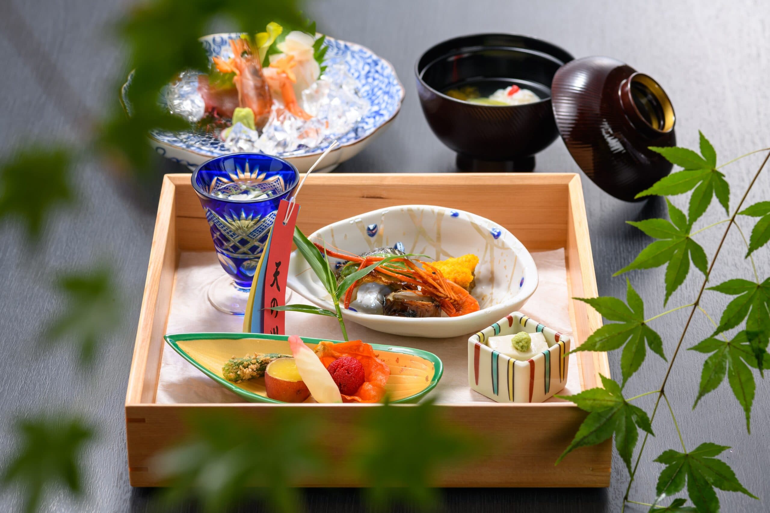 Seasonal kaiseki dinner tray with sashimi and grilled dishes framed by green maple leaves at Kasuga Hotel