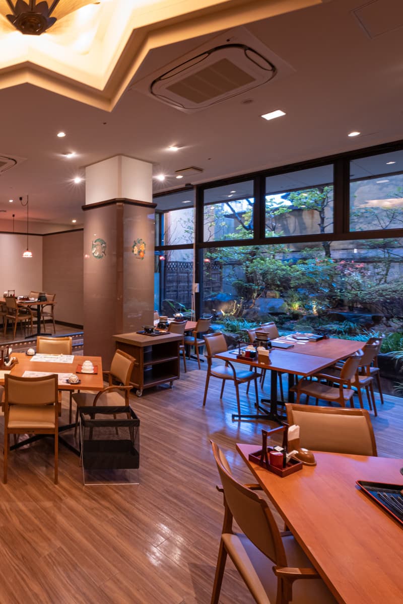 Dining hall at Kinsuikan with set tables beside floor-to-ceiling windows facing a lit garden