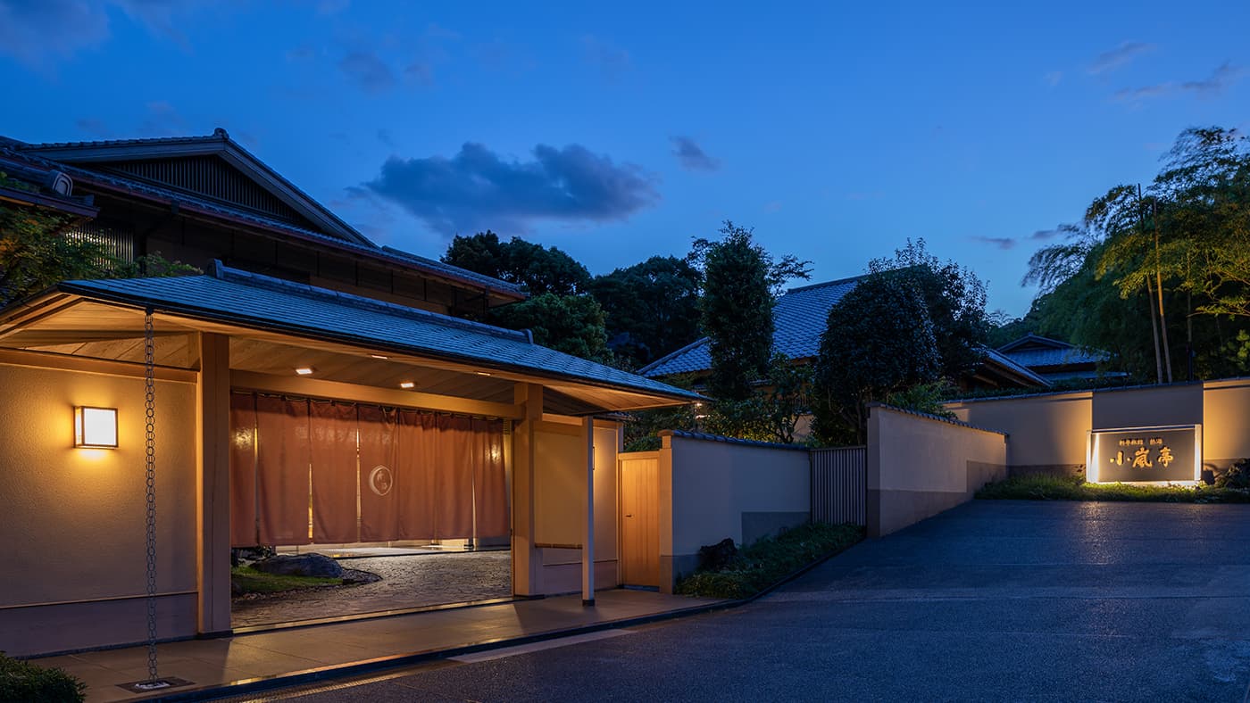 Atami Koarashitei ryokan entrance gate and noren glowing at dusk with a lantern-lit driveway in Atami Onsen