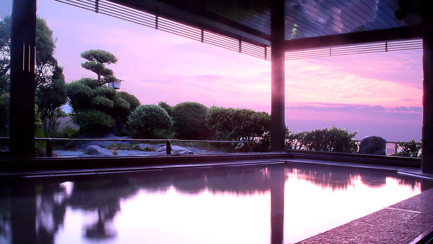 Open-air hot spring bath at dusk reflecting a pink sky with garden pine and distant sea at Atami Koarashitei