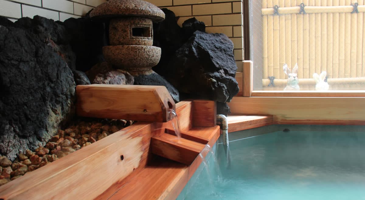 Hot spring source flowing through a cedar spout into a milky bath beside a stone lantern and lava rocks