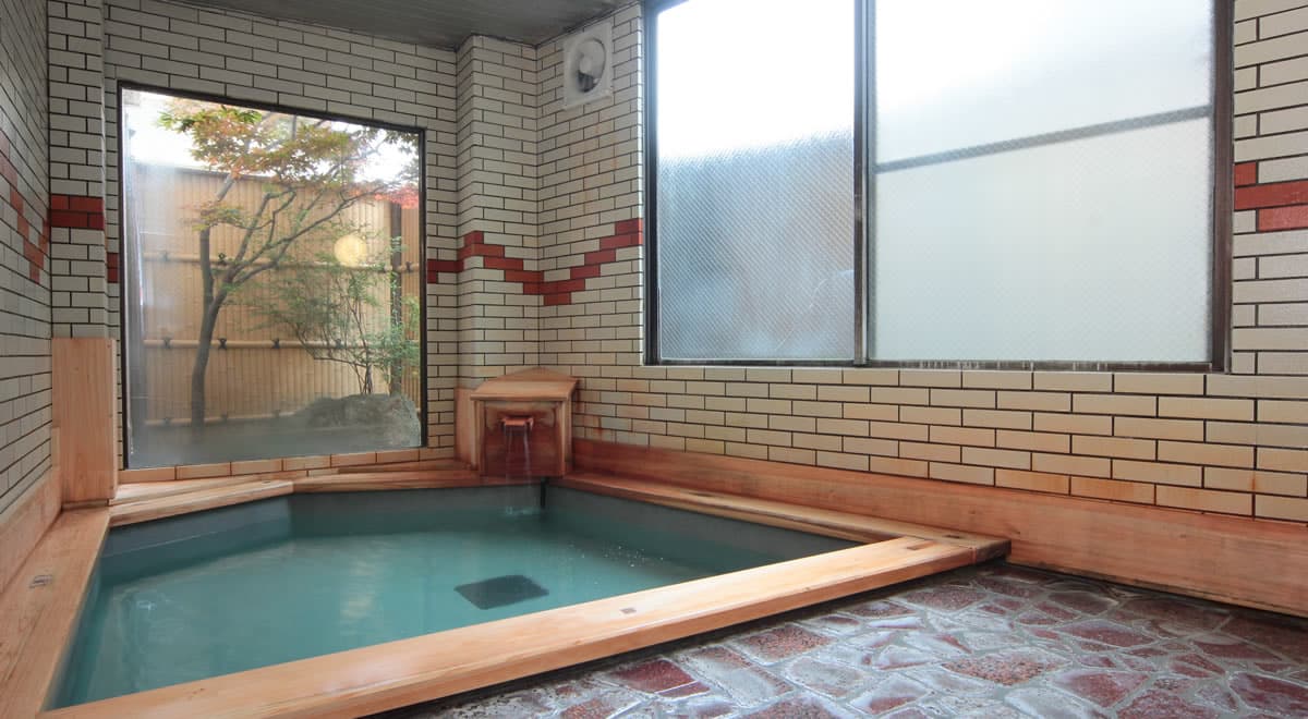 Tiled indoor hot spring bath framed with cedar wood, a small window opening onto a bamboo-fenced garden