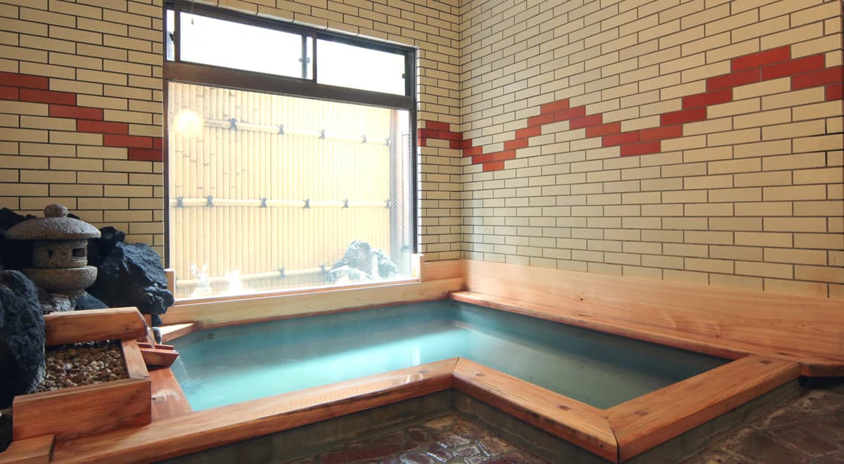 Wide view of a wood-rimmed indoor onsen with a stone lantern and lava rock corner under retro tilework