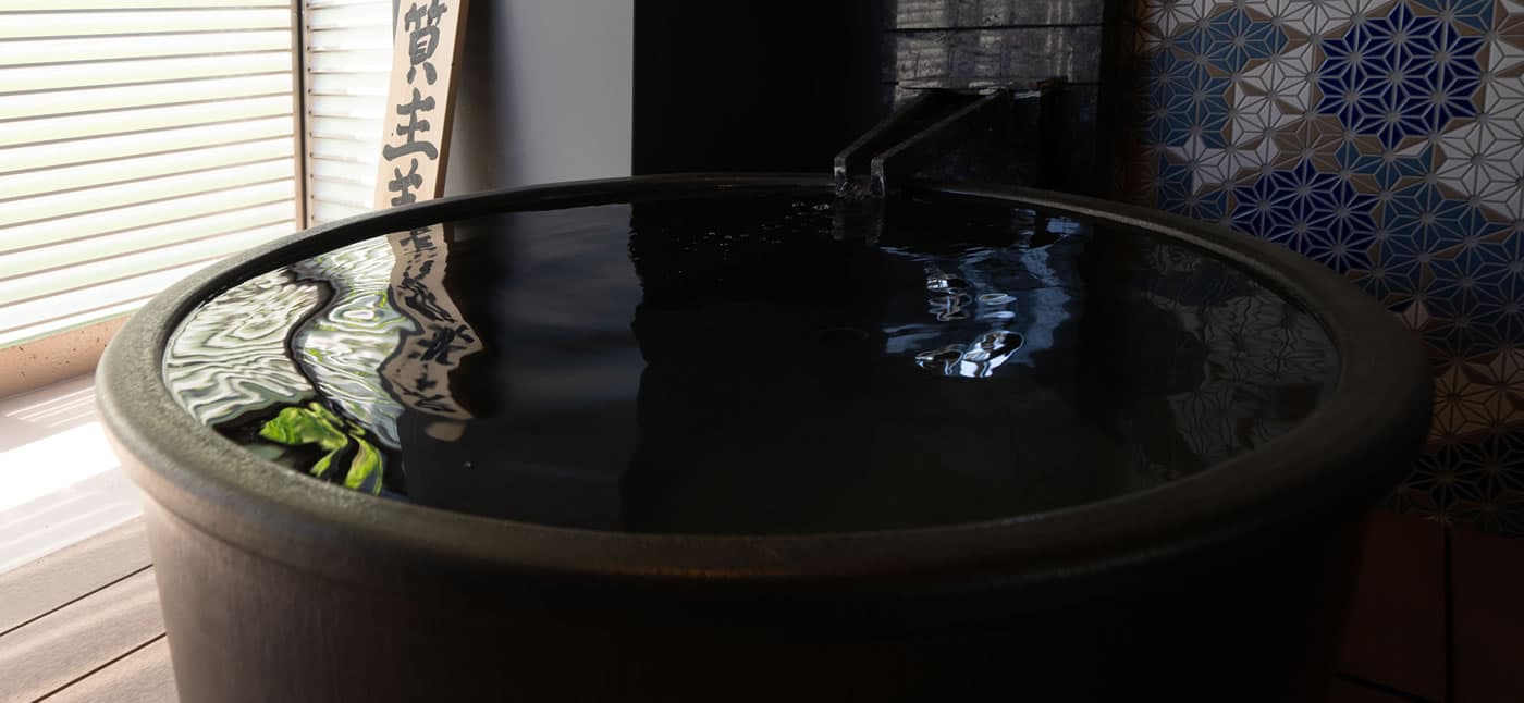 Yubatake Sensui in-room private onsen, a round black ceramic tub with water flowing from a stone spout beside a blue asanoha-tiled wall
