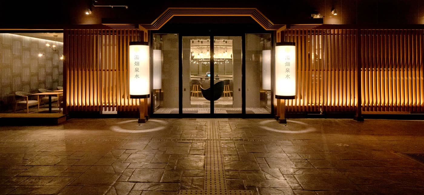 Kusatsu Onsen Yubatake Sensui entrance at night, glass doors flanked by tall paper lanterns reading Yubatake Sensui amid warm-lit timber-slat walls
