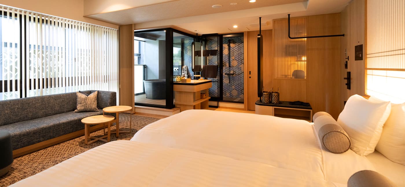 Yubatake Sensui luxury guest room with a low double bed, grey sofa and a glass-walled private bath alcove with a black stone tub