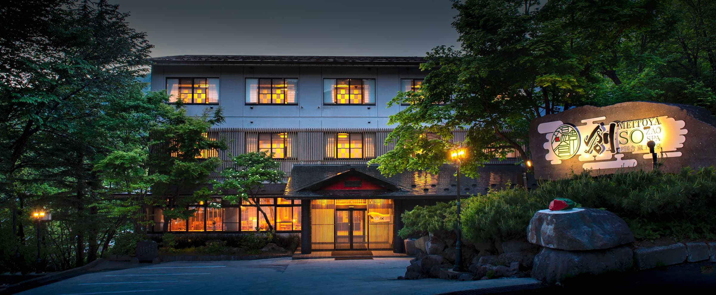 Meitoya So ryokan exterior at dusk in Zao Onsen, warm-lit windows and a stone-lantern approach amid pines