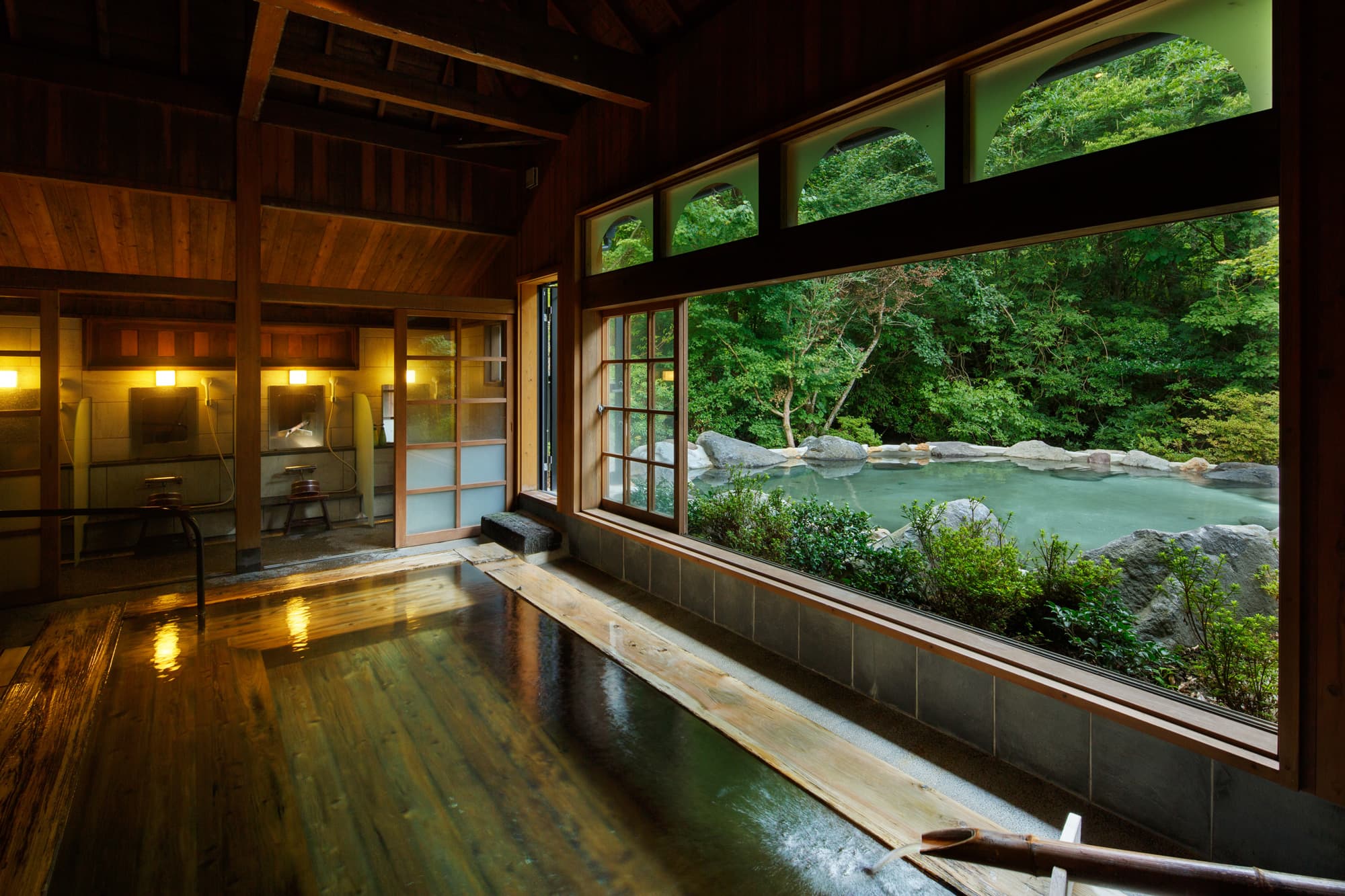 Indoor wood-floored onsen bath with a large window framing green forest and an outdoor stone bath beyond