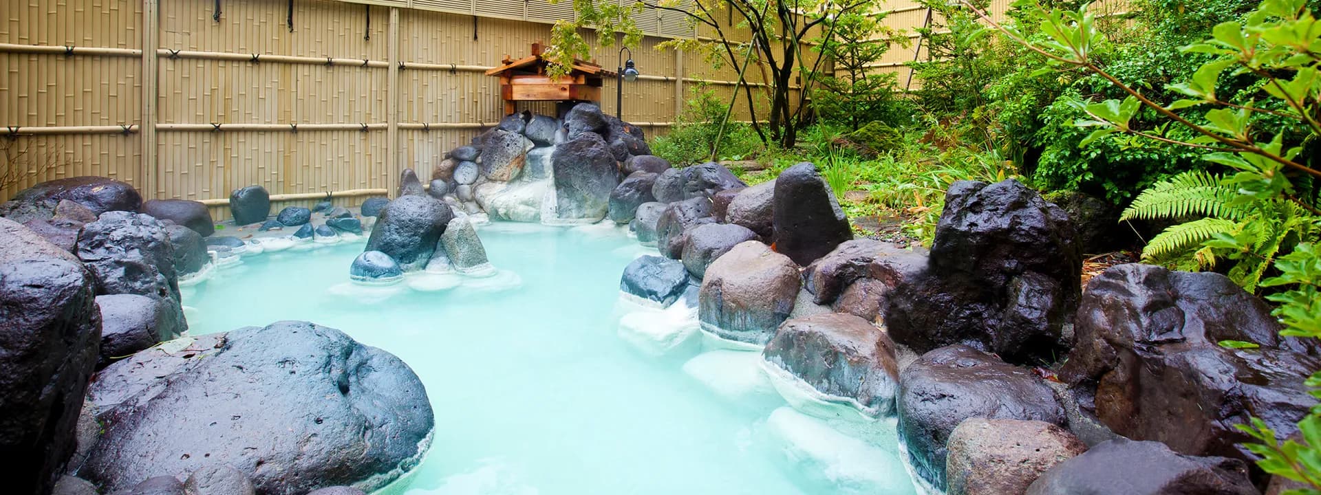 Outdoor rotenburo with milky white onsen water among lava boulders and greenery behind a bamboo fence at Oku Nikko Mori no Hotel