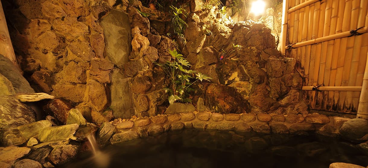 Outdoor rock rotenburo bath at Morizuya at night, with rugged volcanic stone wall, bamboo fence and plantings