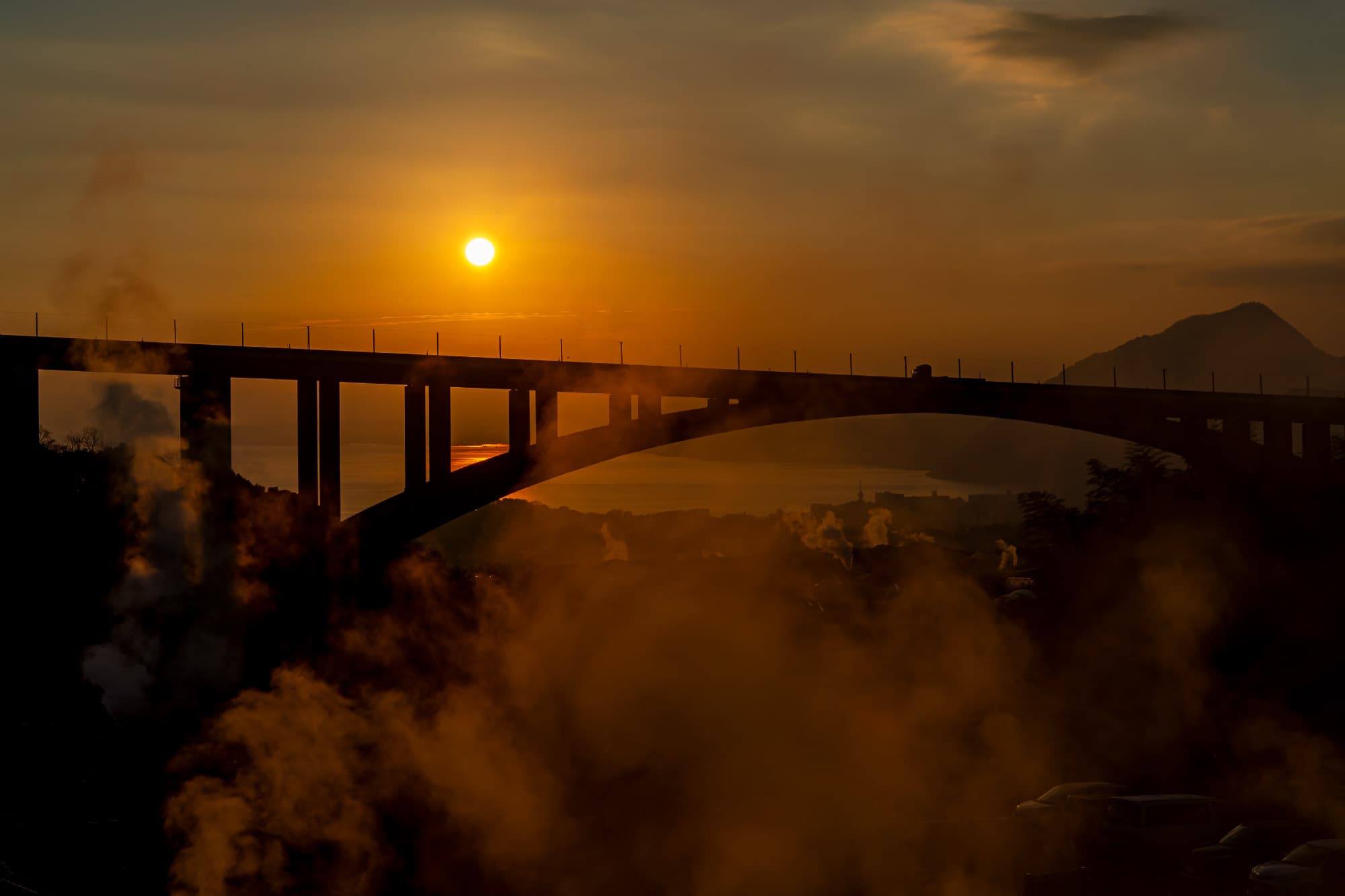 Arched bridge silhouetted against a golden sunset over Beppu bay amid rising hot-spring steam, Okamotoya Ryokan, Myoban Onsen, Beppu