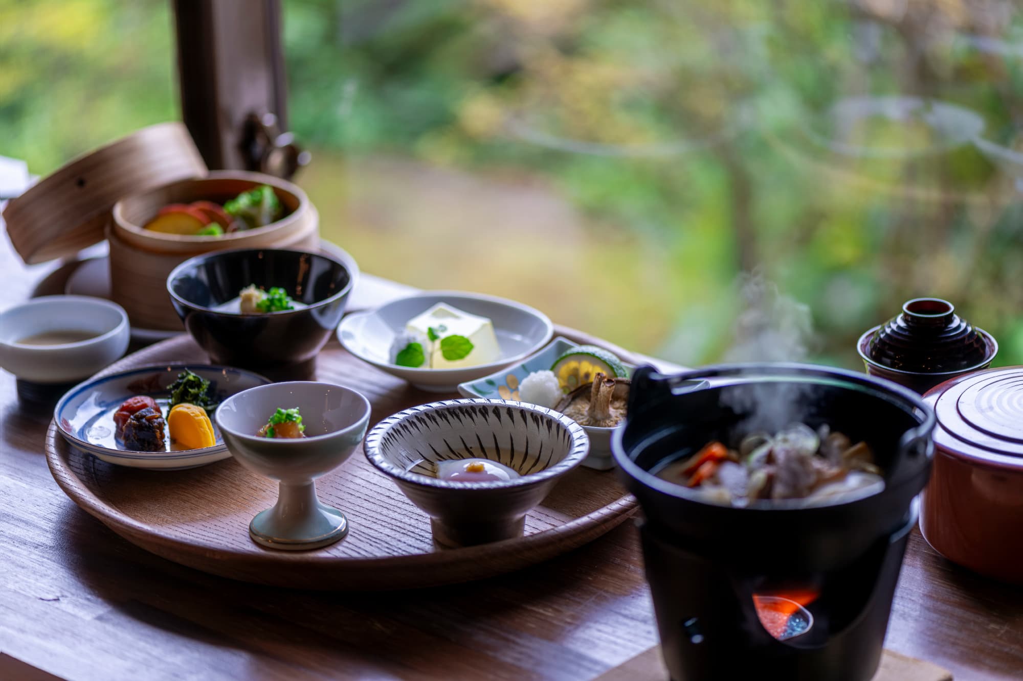 Kaiseki tray of small seasonal dishes and a steaming hotpot beside a window framing autumn foliage, Okamotoya Ryokan, Myoban Onsen, Beppu