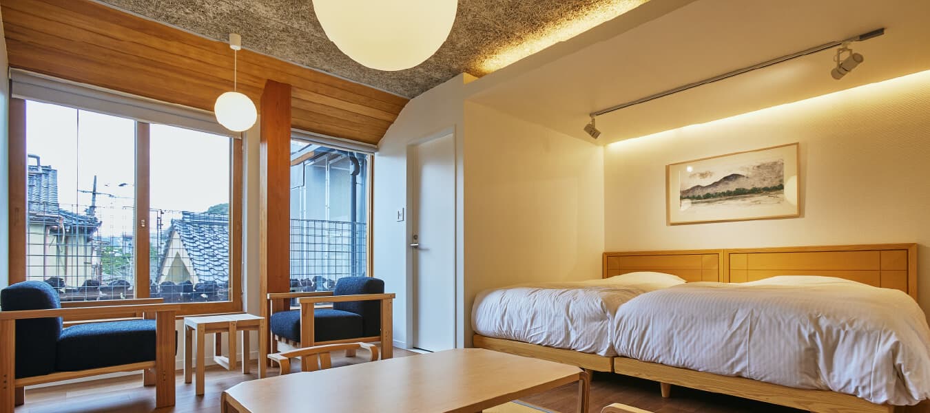 Twin-bed guest room at Tsuchiya with timber ceiling, lounge chairs and a window overlooking the Kinosaki townscape