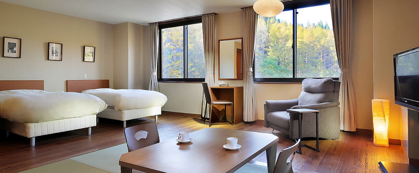 Twin bedroom with armchair and writing desk overlooking autumn-tinted forest through large windows at Ryu Resort and Spa