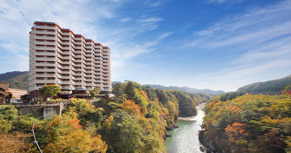 Hotel Sunshine Kinugawa high-rise rising above the Kinugawa River gorge framed by autumn foliage and blue sky at Kinugawa Onsen, Nikko