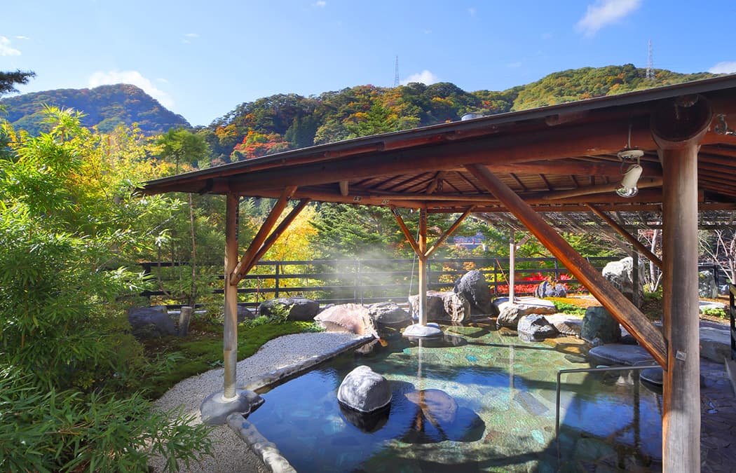 Open-air hot-spring bath surrounded by greenery and hills on a sunny day