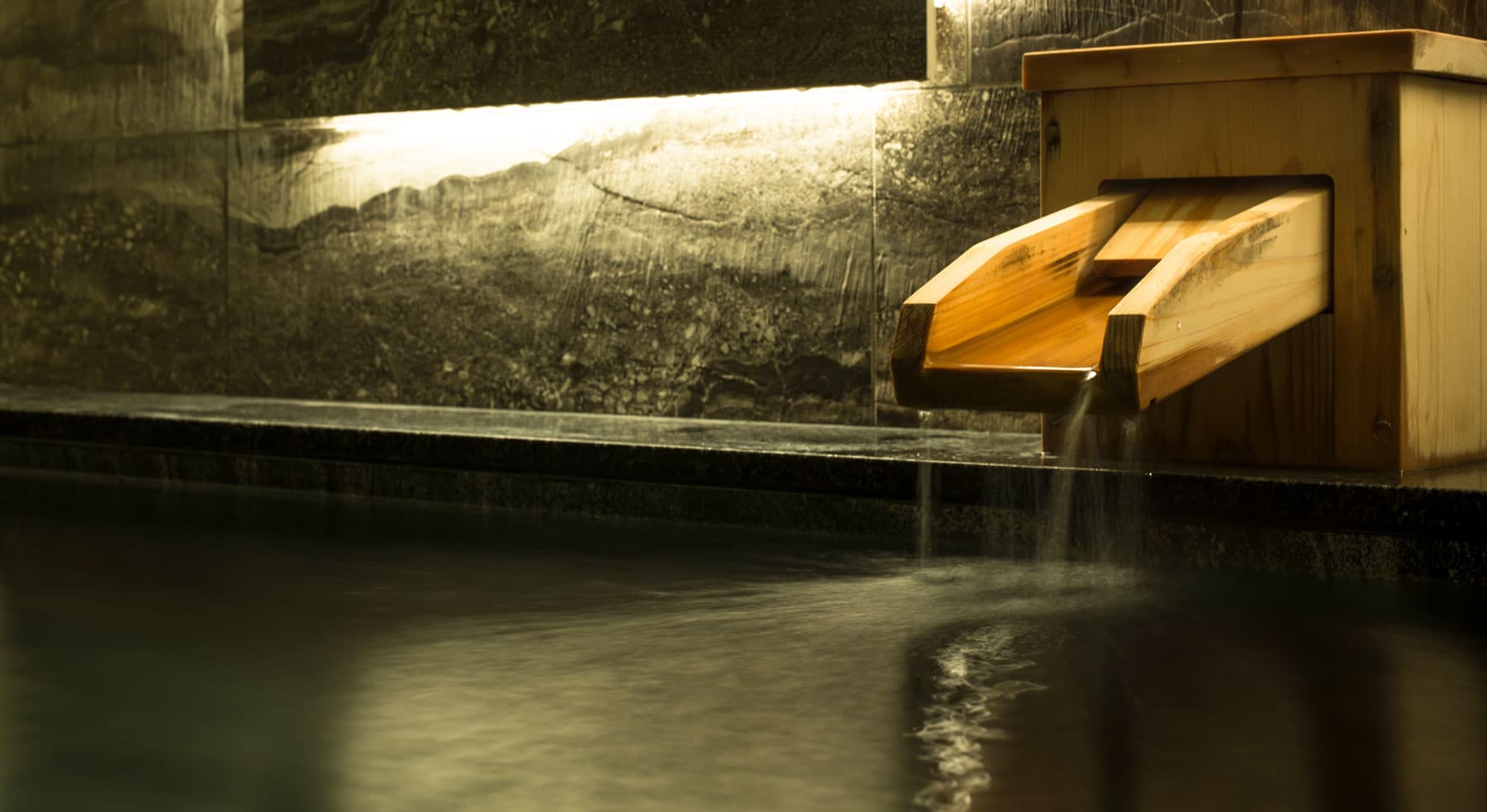 Close-up of the cedar water spout pouring into a dark stone bath at Tokitotoki