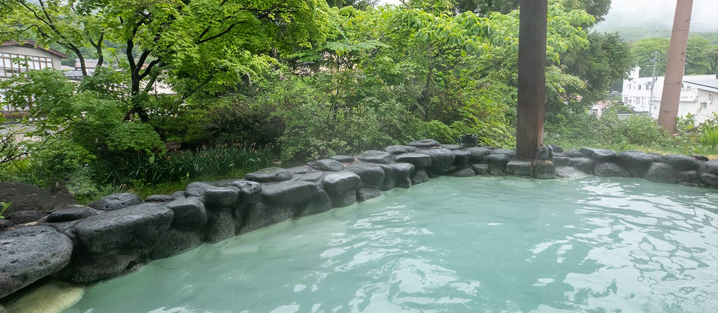 Stone-rimmed outdoor sulfur bath surrounded by fresh green foliage in summer at Zao Onsen