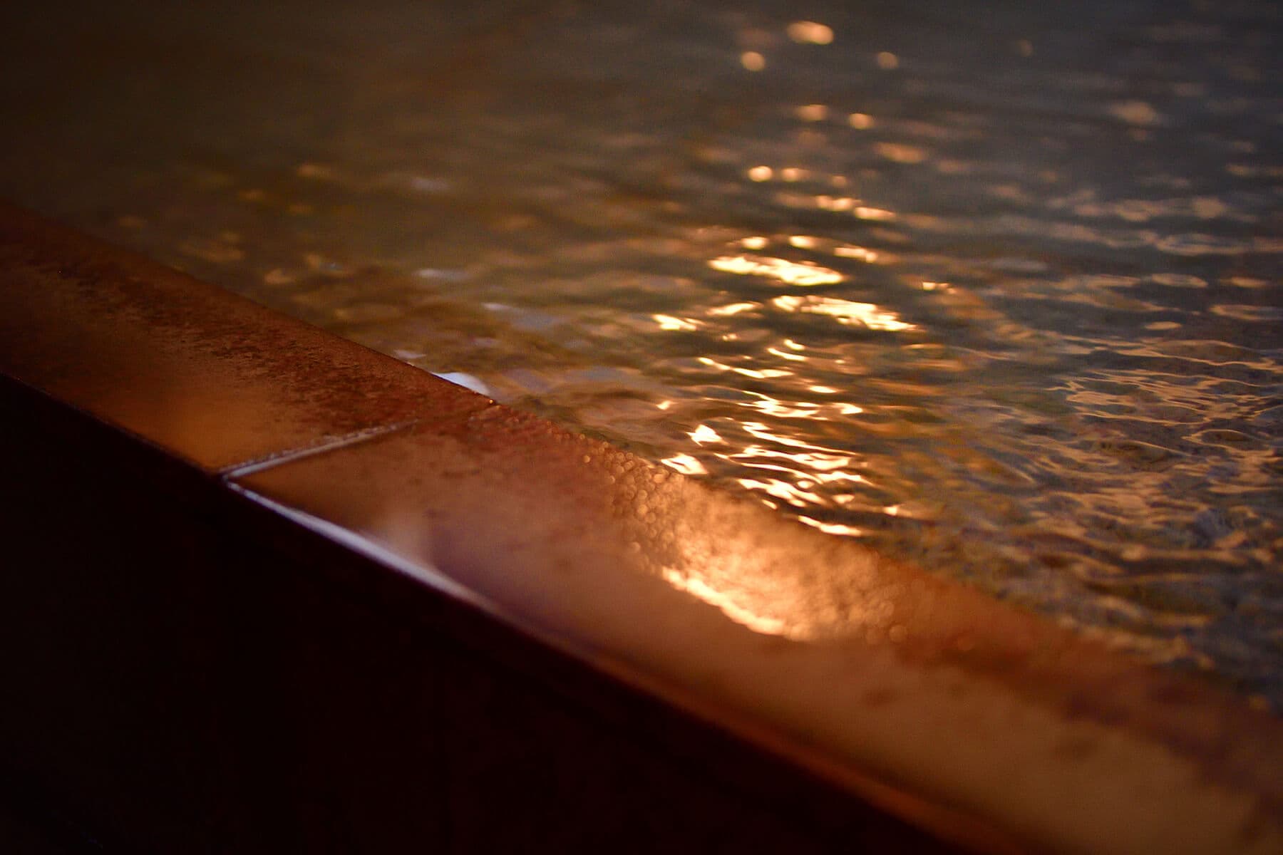 Warm light reflecting on the rippling surface of an indoor onsen bath and its stone edge
